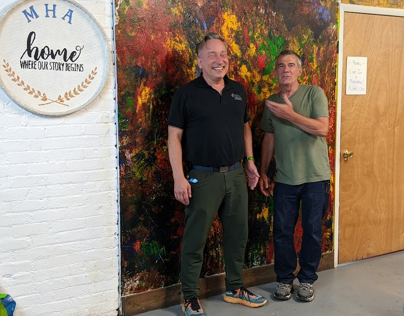 Executive Director Steven Cobb and artist Michael Cain share a laugh in front of the wall mural Cain painted for the Mental Health Association in Chautauqua County. Cain titled his work “Lost in My Rainbow.”